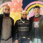 Three men dressed in formal attire standing together, showcasing a variety of styles and accessories, with a colorful balloon backdrop.