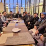 A group of men seated at a long table in a well-lit indoor setting, engaged in discussion, showcasing diverse attire and expressions.